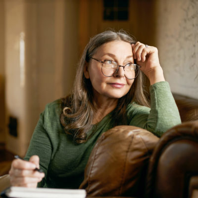 Stylish inspired middle aged woman writer wearing glasses sitting in leather couch with pen making notes. Attractive mature female artist sketching or teacher preparing tasks for online lessons